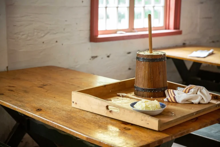 A wooden tray with a plate stacked with butter, a tea towel and butter churn sits a wooden table situated inside of a historical building.