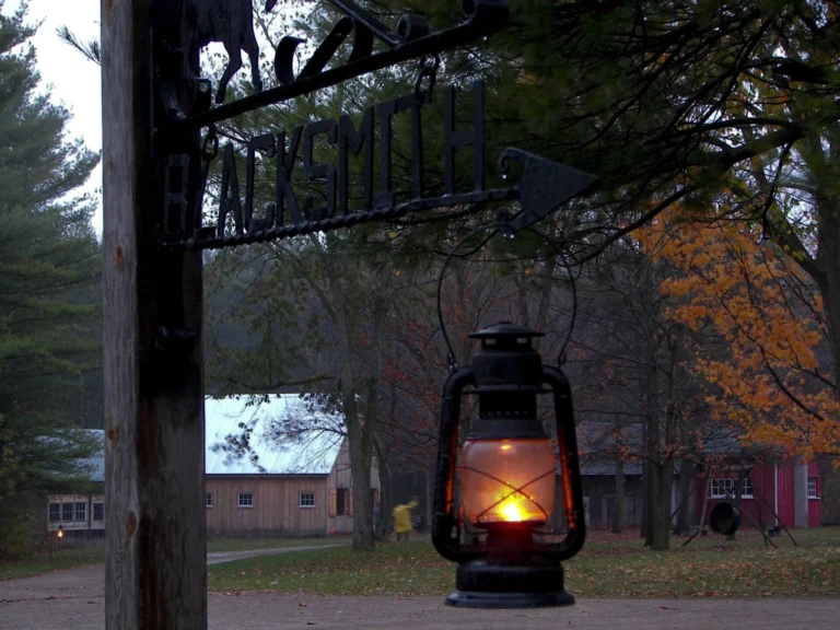 A glowing lantern hangs on sign with the word 'blacksmith'. It is dusk and historical buildings are in the background.